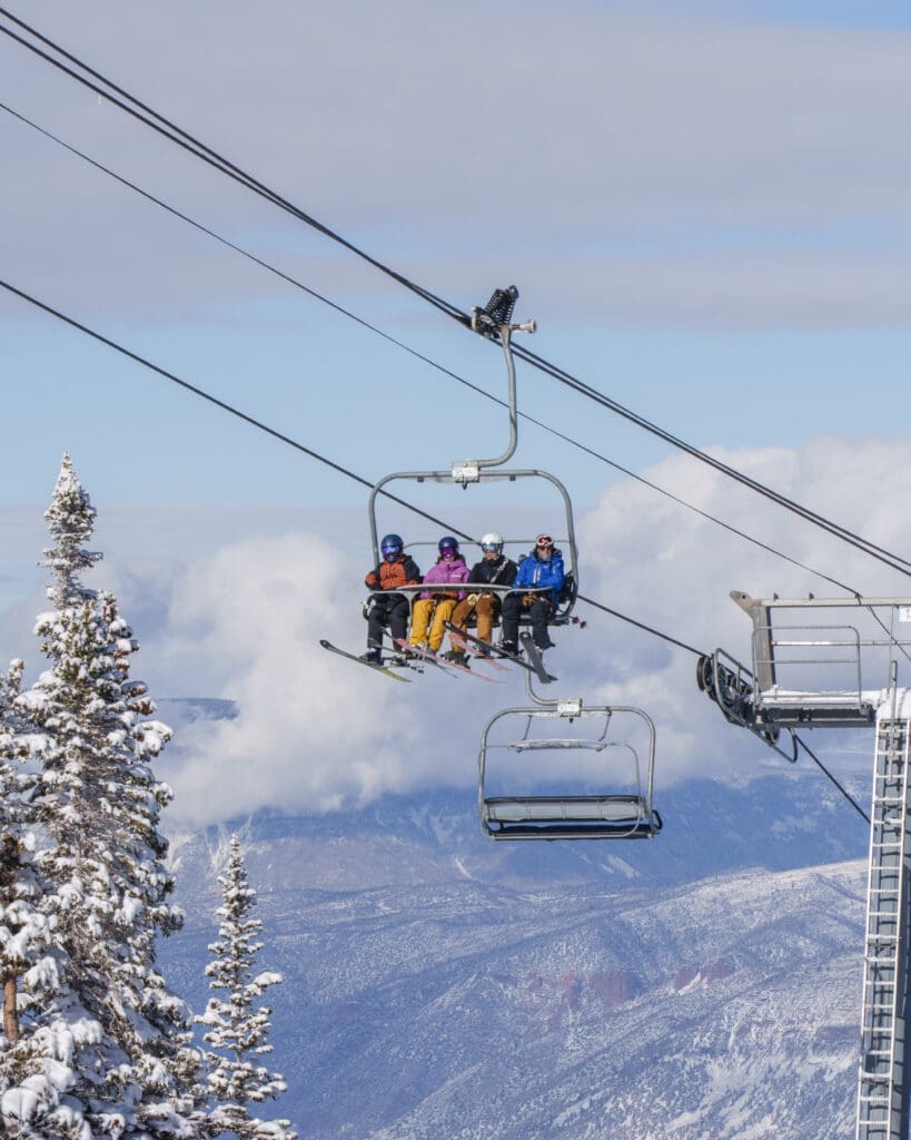 Four skiers riding a chairlift above snowy mountain terrain at Aspen Snowmass with wide alpine views and winter clouds in the distance.
