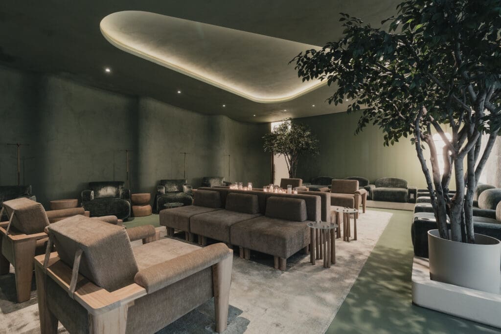 A softly lit wellness lounge at LIVV Natural in Cardiff-by-the-Sea, California, featuring green walls, modern upholstered seating, indoor trees and warm ambient lighting.