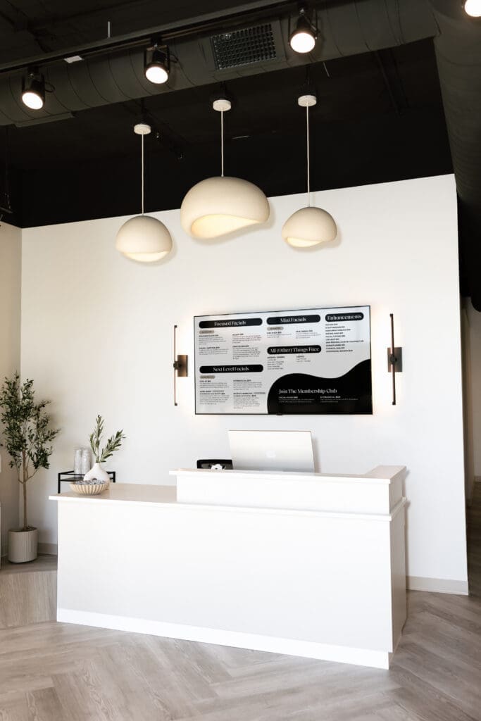 Minimal white reception desk at FACE FOUNDRIÉ Gilbert in SanTan Village featuring modern pendant lighting, a facial treatment menu board and clean contemporary design.