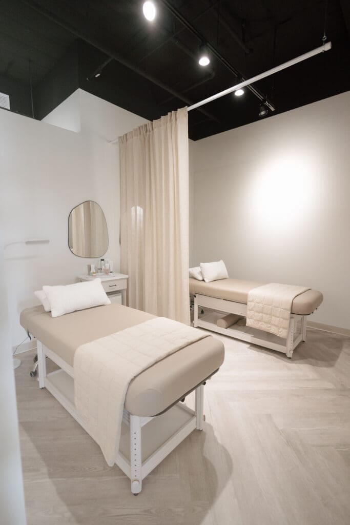 Bright treatment room at FACE FOUNDRIÉ Gilbert with two neutral facial beds, soft lighting and curtain divider for private skincare services in Gilbert Arizona.