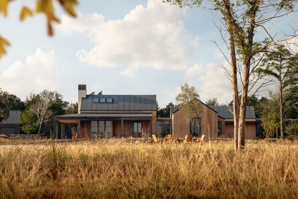 Exterior view of Hideaway Round Top farmhouse in Texas featuring modern wood-clad buildings with metal roofs, expansive porch, outdoor seating and tall golden grasses in the foreground under a partly cloudy sky.