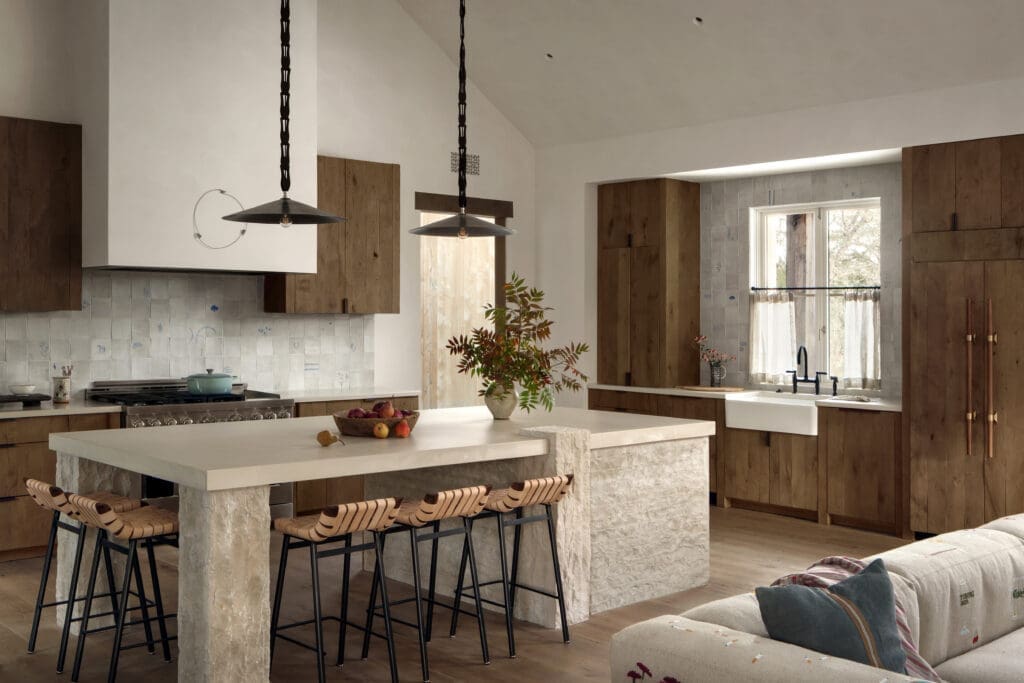 Chef’s kitchen at Hideaway Round Top with large stone island, wood cabinetry, farmhouse sink, pendant lighting, tiled backsplash and bar seating in a warm, neutral-toned interior.