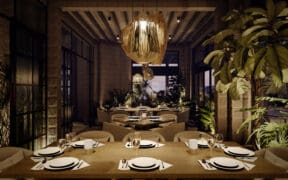 Interior dining room at Flying V Restaurant featuring wood tables set with white plates and wine glasses, woven statement chandeliers, stone walls and abundant indoor greenery under soft evening lighting.