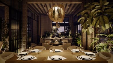 Interior dining room at Flying V Restaurant featuring wood tables set with white plates and wine glasses, woven statement chandeliers, stone walls and abundant indoor greenery under soft evening lighting.