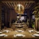 Interior dining room at Flying V Restaurant featuring wood tables set with white plates and wine glasses, woven statement chandeliers, stone walls and abundant indoor greenery under soft evening lighting.
