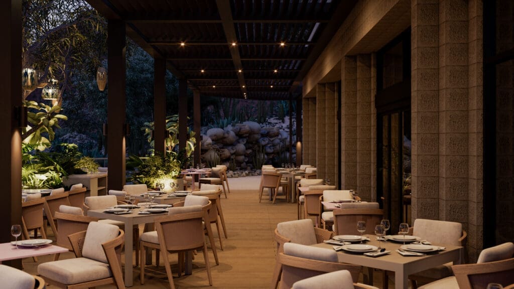 Outdoor dining terrace at Flying V Restaurant with neatly set tables, wooden pergola ceiling, soft lighting and desert landscape with rocks and cacti visible beyond the patio at night.
