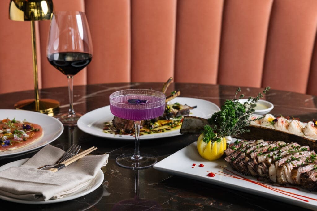Steak sliced and plated with lemon garnish alongside sushi, small plates, a purple cocktail and a glass of red wine at JING Scottsdale.