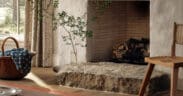 Interior of Hideaway Round Top farmhouse featuring a plaster fireplace with brick interior, natural stone hearth, wooden flooring, woven basket with blue textile, striped curtains and a simple wooden chair in a warm, neutral-toned living space.