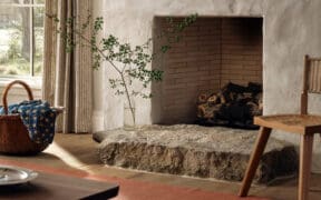 Interior of Hideaway Round Top farmhouse featuring a plaster fireplace with brick interior, natural stone hearth, wooden flooring, woven basket with blue textile, striped curtains and a simple wooden chair in a warm, neutral-toned living space.