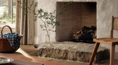 Interior of Hideaway Round Top farmhouse featuring a plaster fireplace with brick interior, natural stone hearth, wooden flooring, woven basket with blue textile, striped curtains and a simple wooden chair in a warm, neutral-toned living space.