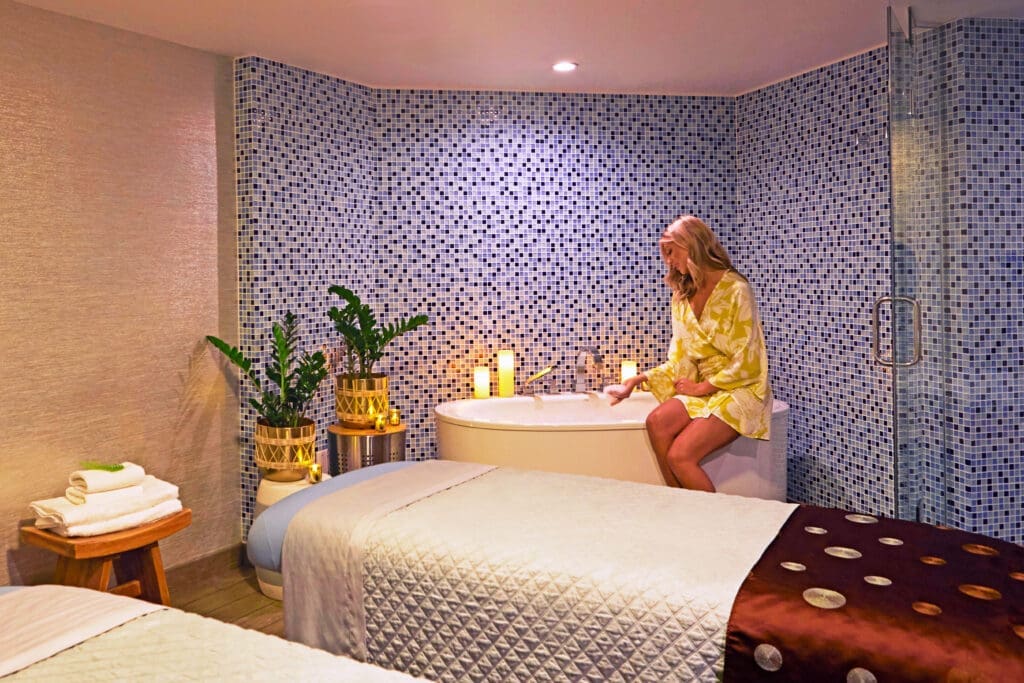 A softly lit treatment room at SpaTerre at Kona Kai San Diego featuring two massage tables, blue mosaic tile walls and a soaking tub with candles.
