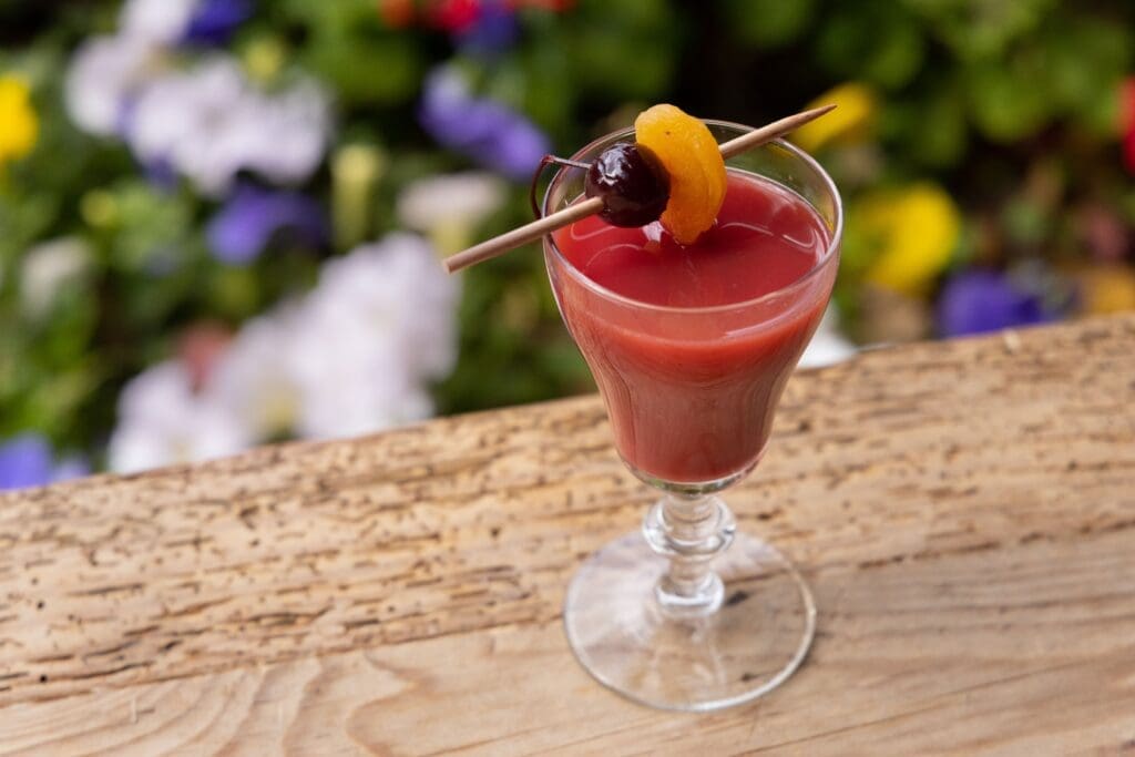 Signature cocktail from v garnished with fruit and served against a garden backdrop.