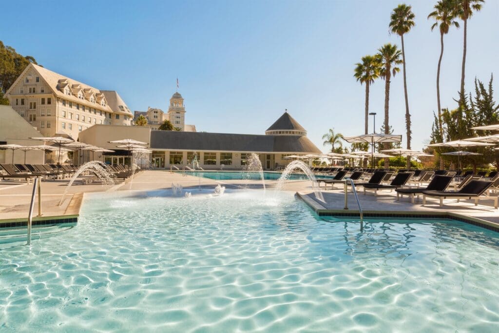 The main pool at Claremont Resort & Club in Berkeley, California, featuring clear turquoise water with arched fountains, palm trees, lounge chairs and the historic white resort building overlooking the San Francisco Bay.