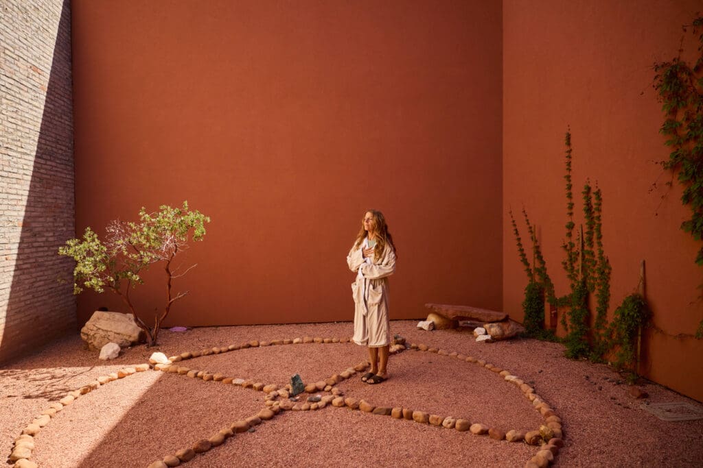 A guest in a robe stands inside a circular stone labyrinth courtyard at Mii amo in Sedona, Arizona, surrounded by terracotta walls, desert landscaping and soft natural light.