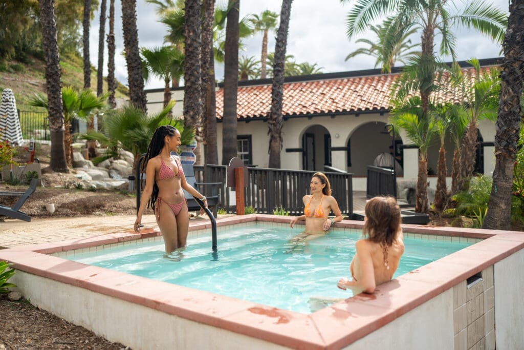 Guests enjoying a geothermal soaking pool at Murrieta Hot Springs Resort in California surrounded by palm trees and Spanish-style architecture. 