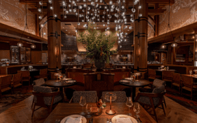 Interior of Napa Rose at Disney’s Grand Californian Hotel & Spa featuring wood columns, round dining tables, leather seating, open kitchen and a chandelier with suspended glass orbs.