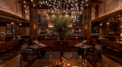 Interior of Napa Rose at Disney’s Grand Californian Hotel & Spa featuring wood columns, round dining tables, leather seating, open kitchen and a chandelier with suspended glass orbs.