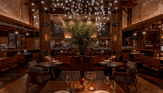 Interior of Napa Rose at Disney’s Grand Californian Hotel & Spa featuring wood columns, round dining tables, leather seating, open kitchen and a chandelier with suspended glass orbs.