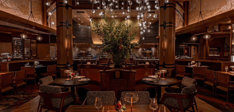 Interior of Napa Rose at Disney’s Grand Californian Hotel & Spa featuring wood columns, round dining tables, leather seating, open kitchen and a chandelier with suspended glass orbs.
