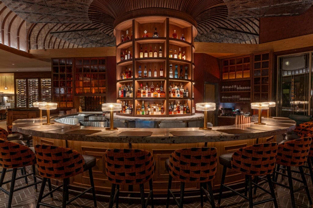 Bar area at Napa Rose with circular marble bar top, illuminated liquor shelves, patterned bar stools and warm wood paneling under decorative ceiling details.