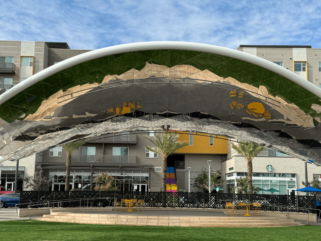 Curved canopy stage at Novus Place in Tempe with green lawn, palm trees and surrounding retail buildings.