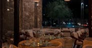 Interior dining room at Santo Agave in Peoria featuring woven pendant lights, stone accent walls, glass-topped tables, upholstered chairs and an open patio setting at night.