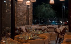 Interior dining room at Santo Agave in Peoria featuring woven pendant lights, stone accent walls, glass-topped tables, upholstered chairs and an open patio setting at night.