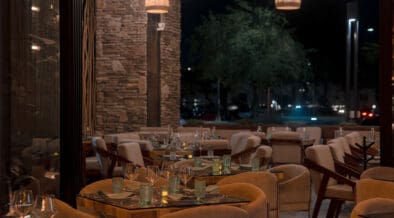 Interior dining room at Santo Agave in Peoria featuring woven pendant lights, stone accent walls, glass-topped tables, upholstered chairs and an open patio setting at night.