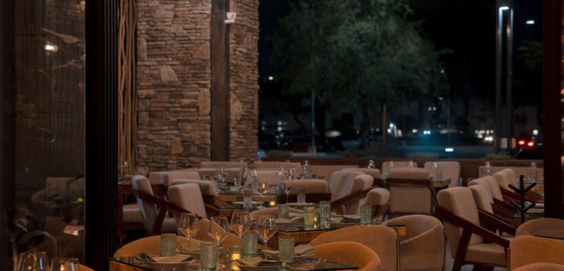 Interior dining room at Santo Agave in Peoria featuring woven pendant lights, stone accent walls, glass-topped tables, upholstered chairs and an open patio setting at night.