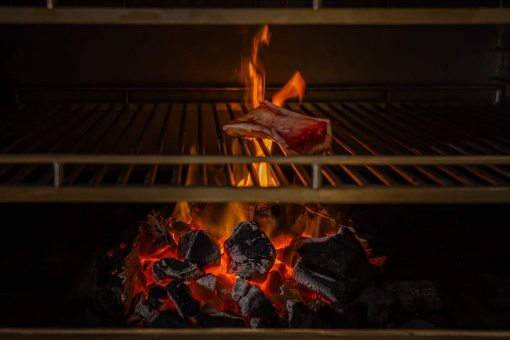 Steak cooking over open flame inside a Josper charcoal oven at Santo Agave, with glowing coals and visible flames beneath the grill.