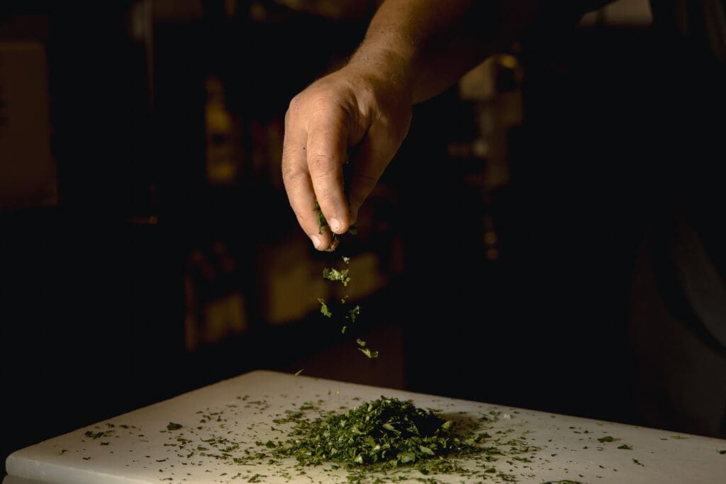Chef’s hand sprinkling fresh chopped herbs over a cutting board in a dimly lit kitchen at Santo Agave in Peoria.