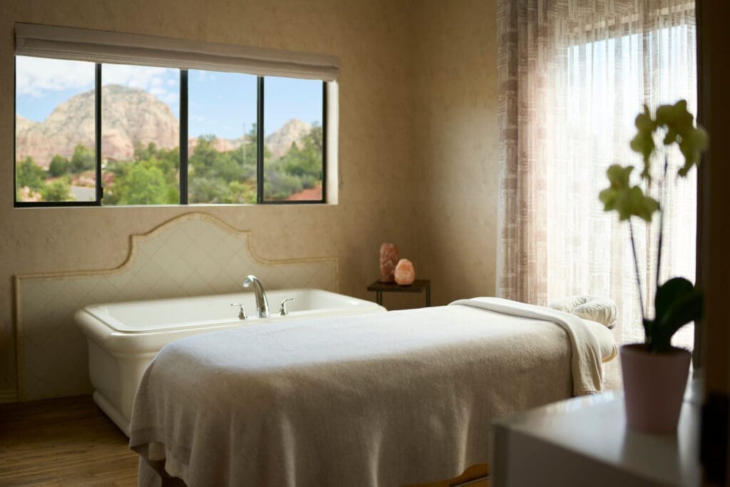 A spa treatment room at The Wilde Resort & Spa in Sedona, Arizona, featuring a massage table, soaking tub, soft natural light and a window overlooking red rock formations.
