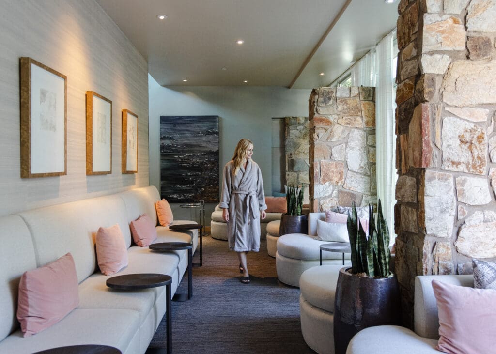 A guest in a robe walks through a softly lit relaxation lounge at Sanctuary Spa in Scottsdale, Arizona, featuring stone walls, neutral seating, pink accent pillows and a calm, modern desert aesthetic.