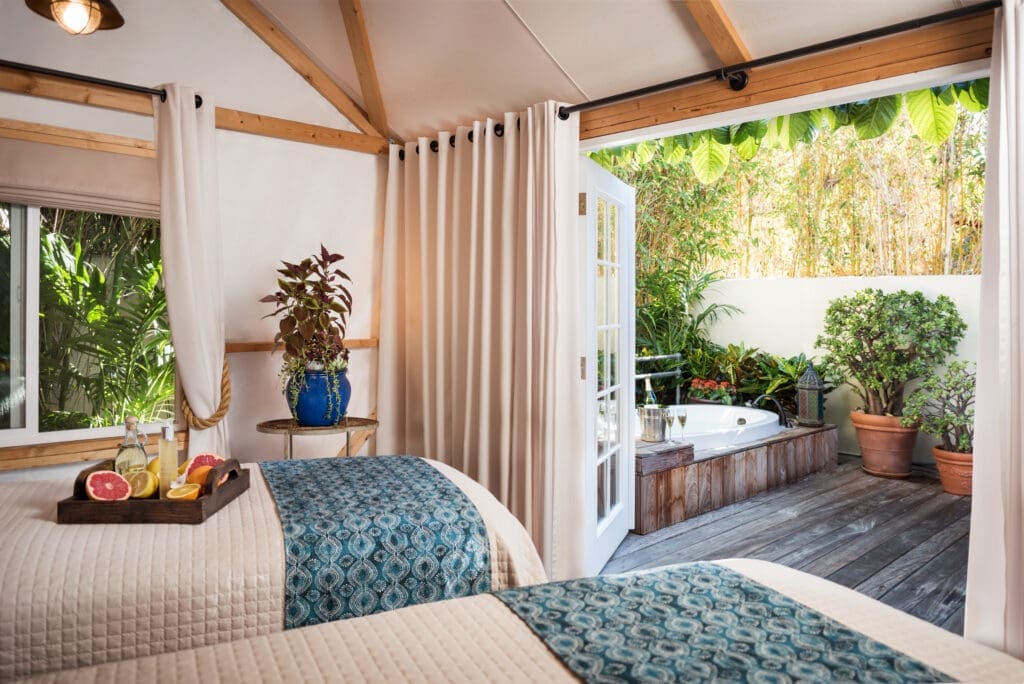 A treatment suite at Spa Estancia in La Jolla, California, with two massage tables and an open doorway leading to a private outdoor soaking tub surrounded by greenery.