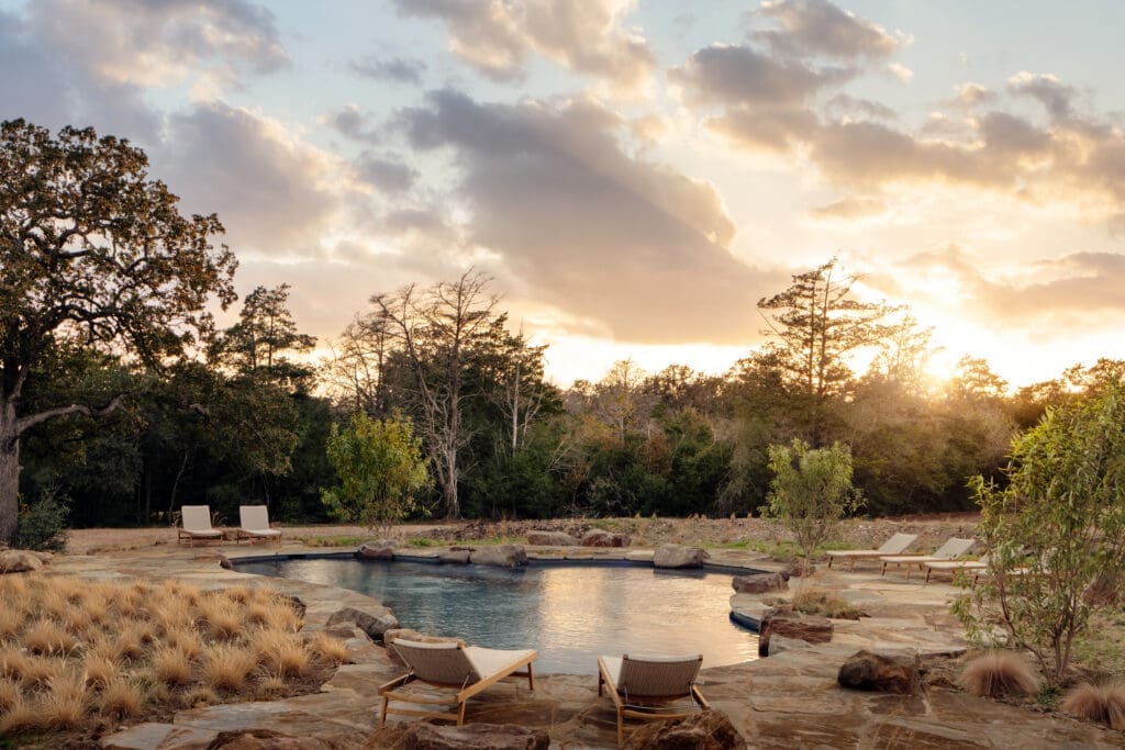 Spring-inspired pool at Hideaway Round Top surrounded by natural stone decking, lounge chairs, native grasses and trees, with a golden sunset sky over the Texas countryside.