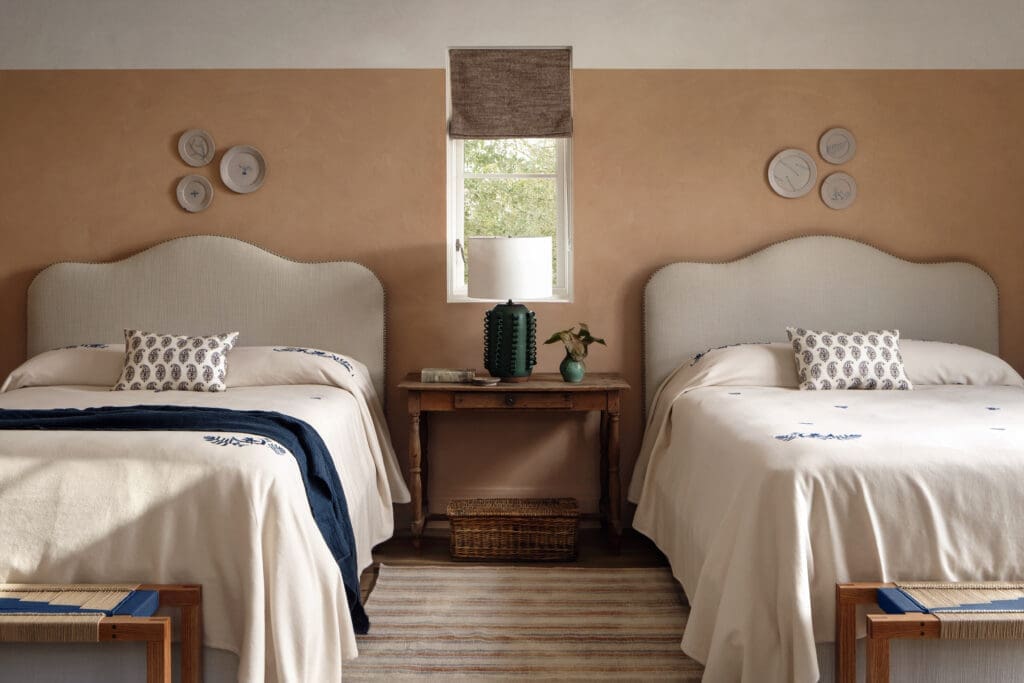 Double queen bedroom at Hideaway Round Top featuring two upholstered headboards, neutral bedding with blue embroidered details, wooden nightstand with lamp, woven rug, soft earth-toned walls and natural light from a centered window.