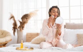 Person enjoying a quiet self-care moment while applying skincare in a softly lit bedroom, reflecting indulgent Valentine’s Day rituals.