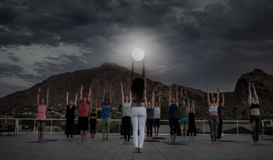 A group of guests practice rooftop yoga at Mountain Shadows Resort Scottsdale in Arizona, standing on mats with arms raised toward the moon, with Camelback Mountain visible in the background at dusk.