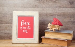 Framed artwork reading “love is in the air” displayed on a wooden surface beside stacked books and a small red heart decoration, representing sentimental Valentine’s Day gifts.