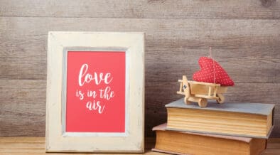 Framed artwork reading “love is in the air” displayed on a wooden surface beside stacked books and a small red heart decoration, representing sentimental Valentine’s Day gifts.
