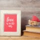 Framed artwork reading “love is in the air” displayed on a wooden surface beside stacked books and a small red heart decoration, representing sentimental Valentine’s Day gifts.