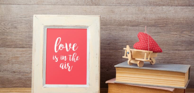 Framed artwork reading “love is in the air” displayed on a wooden surface beside stacked books and a small red heart decoration, representing sentimental Valentine’s Day gifts.