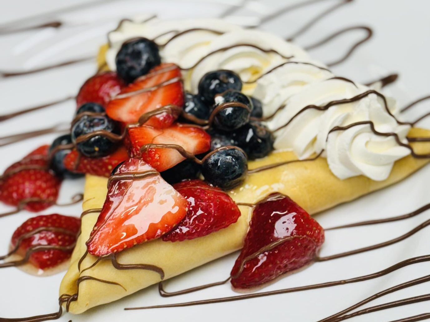 Crepe topped with fresh strawberries, blueberries, whipped cream, and chocolate drizzle on a white plate at A Bite of Belgium.
