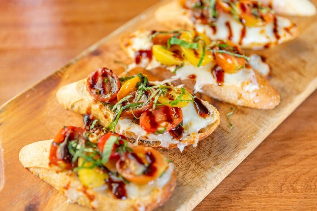 Toasted bruschetta topped with melted cheese, heirloom cherry tomatoes, fresh basil and balsamic glaze on a wooden serving board at Bistro & Wine Bar.