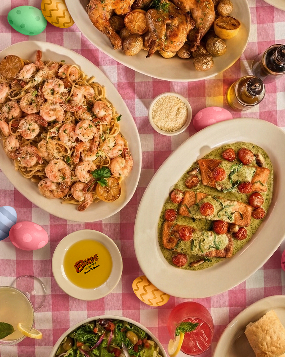 Overhead view of a colorful Easter table with shrimp pasta, roasted chicken, salmon, salad and cocktails on a pink checkered tablecloth with decorative eggs.