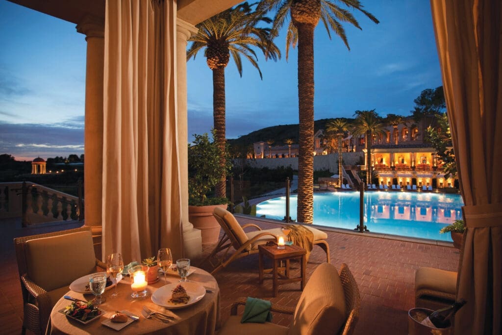 Candlelit outdoor dining table set beside a resort pool at dusk, with palm trees, lounge chairs, and a grand illuminated building in the background.