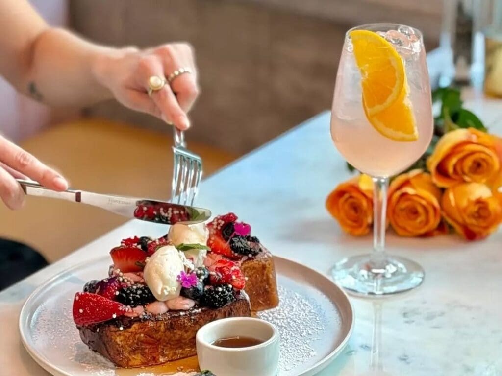 A person cutting into a berry and cream topped French Toast with a cocktail and flowers nearby at Canvas Cafe.
