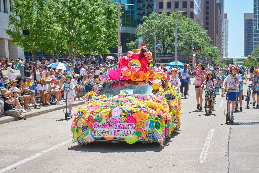 Brightly decorated car covered in colorful flowers and shapes driving down a crowded Houston street with children riding bikes nearby.