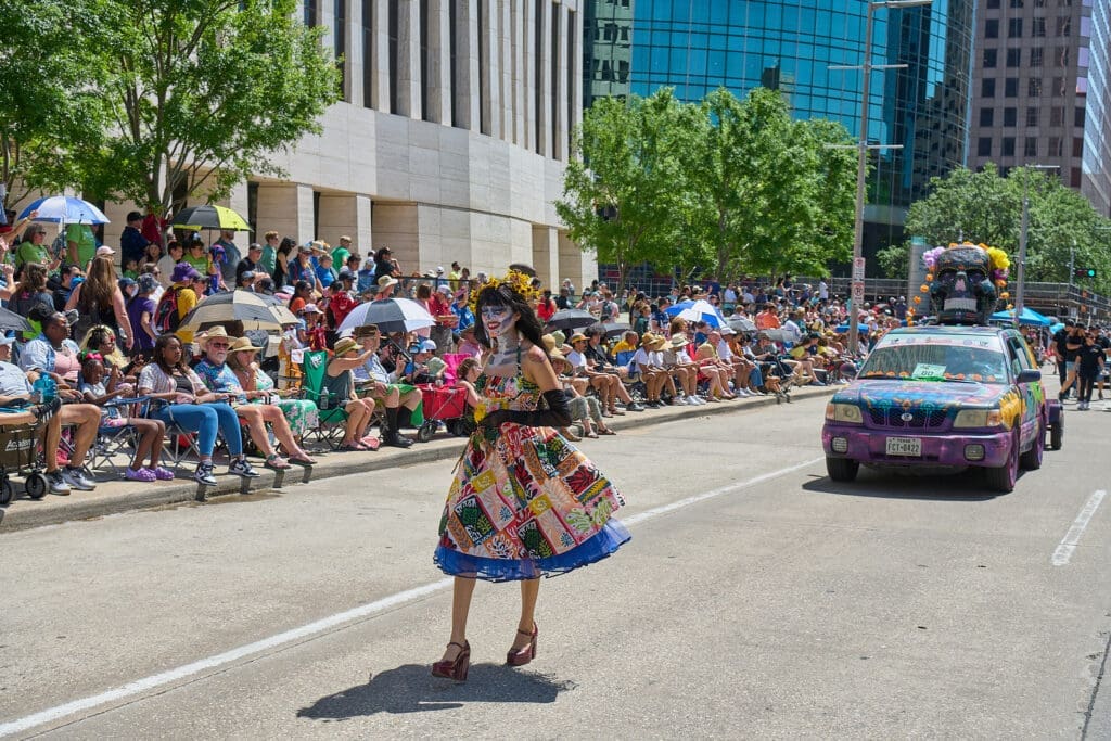 Performer in colorful dress dancing in the street with a decorated car featuring a large skull sculpture behind her.