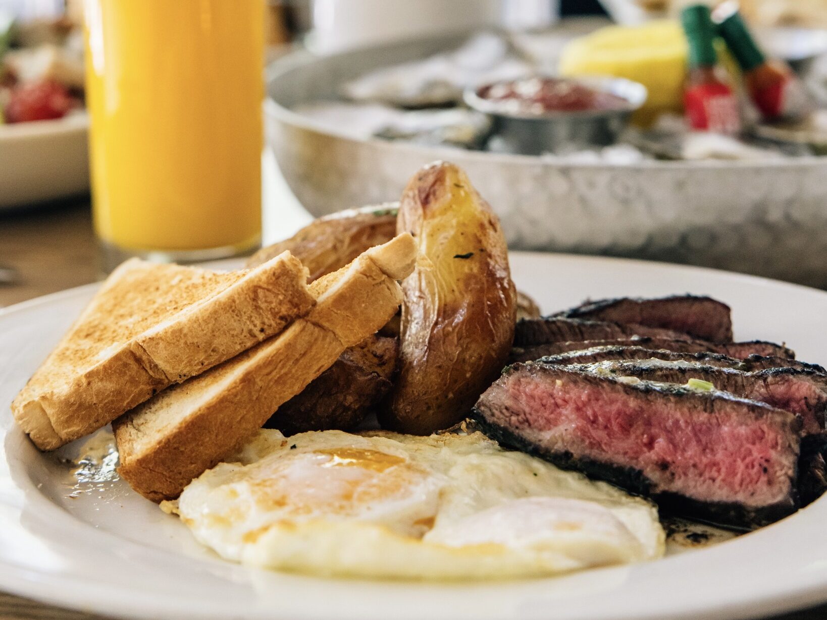 Close-up of a plated brunch dish with steak, fried eggs, roasted potatoes and toast alongside a glass of orange juice at Corrine, Le Méridien Denver Downtown.